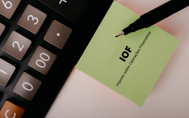 calculadora em cima da mesa ao lado de uma caneta e uma folha de papel verde escrito IOF Imposto sobre Operações Financeiras