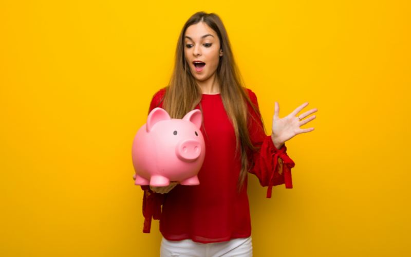 Moça com vestido vermelho sobre a parede amarela surpreendida ao guardar um piggybank mostrando como juntar dinheiro com o desafio das 52 semanas