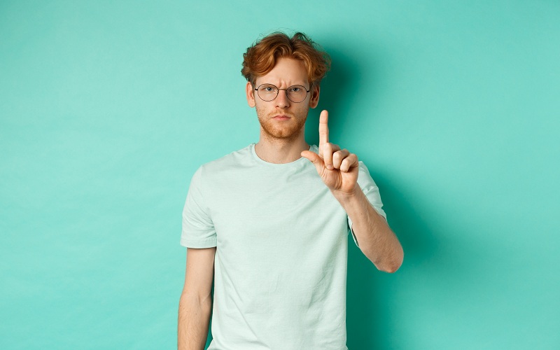 Homem jovem sério e zangado, com cabelo vermelho, usando óculos, mostrando um gesto de pare, dizendo não, balançando o dedo em desaprovação simbolizando os motivos para não emprestar o CPF ou cartão de crédito para terceiros