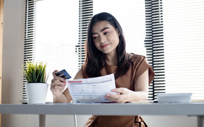Jovem mulher asiática segurando o cartão de crédito e a conta com um sorriso na mesa de trabalho em casa simbolizando a alegria em saber como negociar as dívidas no cartão de crédito