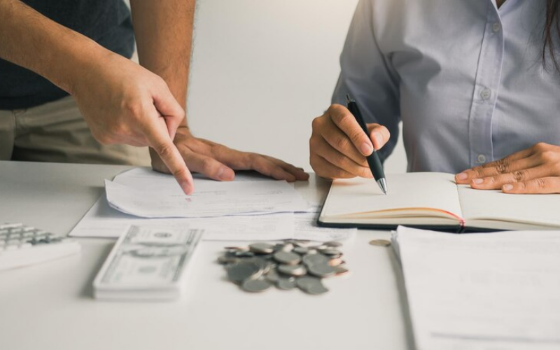 imagem de um casal de homem e mulher ao fundo revendo as finanças para casa, ela com contas no papel e ele apontando para moedas