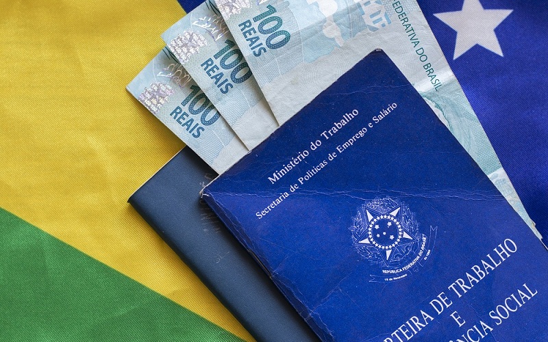 bandeira do Brasil e em cima dela carteira de trabalho com notas de cem reais, simulando o14° salário do INSS será pago em 2022